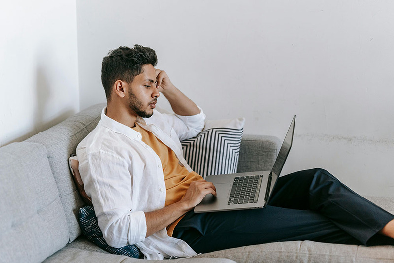 A young man rests on the couch, looking at his laptop as he applies to be an ordained minister online