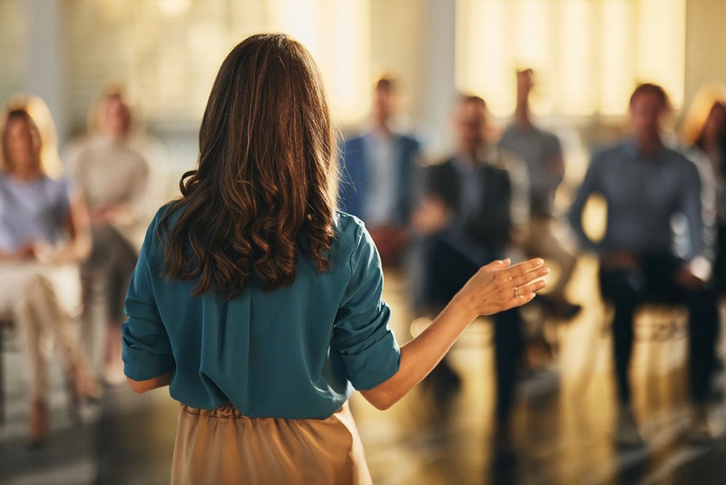 Woman stands with her back to the photographer, she is speaking to a group of people in the background