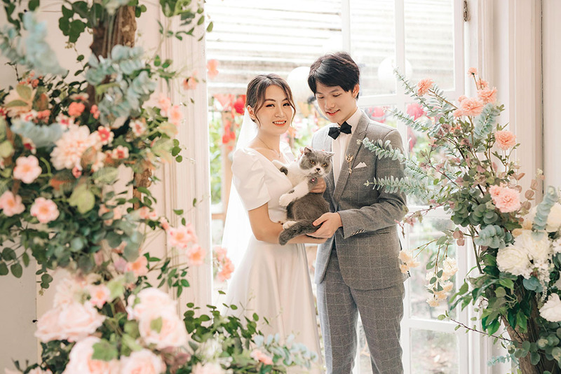 Bride and groom pose with a cat for wedding photos next to elaborate wedding arch with flowers