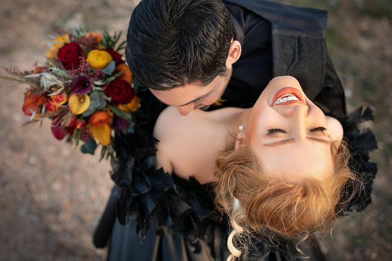 A bride and groom photographed from above as they embrace, laughing and happy on the wedding day. The bride is holding a bouquet of fall wedding flowers, and they are both wearing all black 
