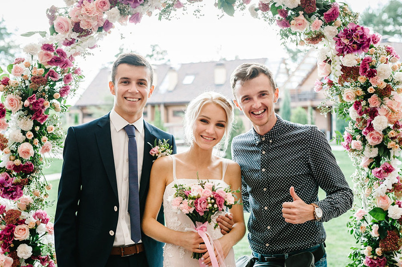 A bride and groom pose with their friend officiant on the wedding day, next to a beautiful wedding arch decorated with roses