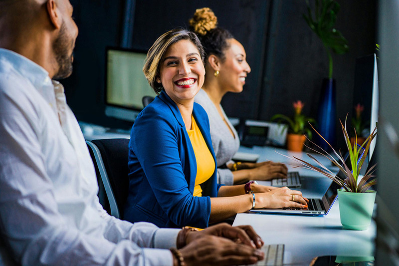 A happy woman smiles while using her laptop, friends sit nearby.