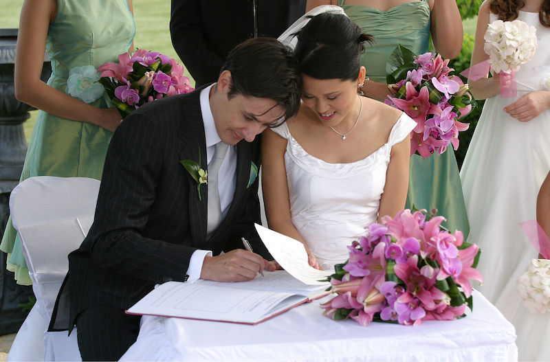 photograph of a young couple at their wedding ceremony, the bride holds a bouquet of flowers while the groom signs the marriage license after the ceremony