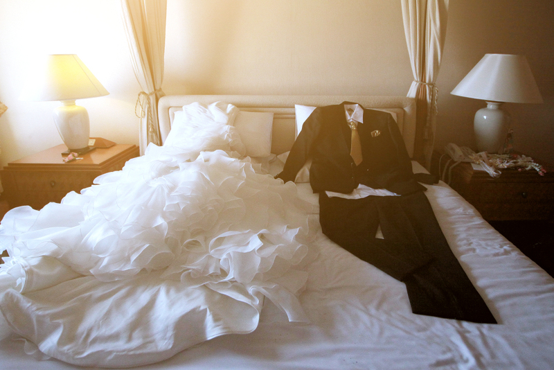 A white wedding dress and suit on a bed as a couple chooses their wedding attire.