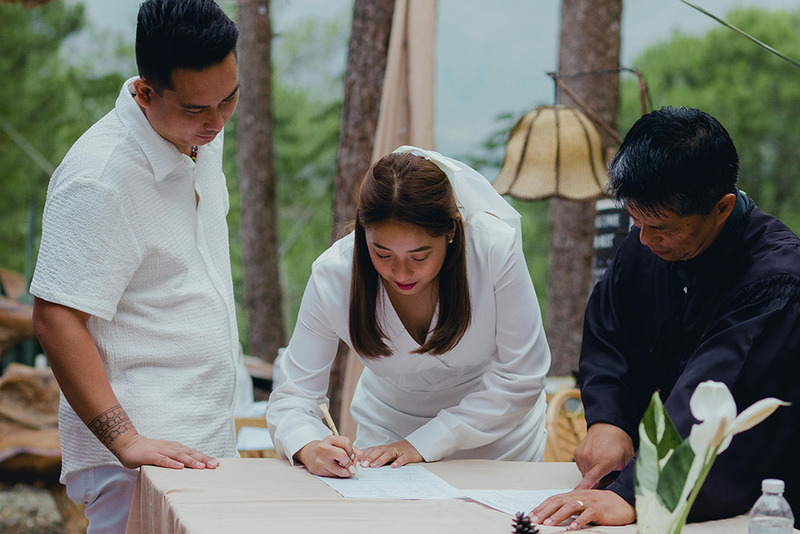 Bride signs marriage license next to groom while the wedding officiant watches after outdoor wedding