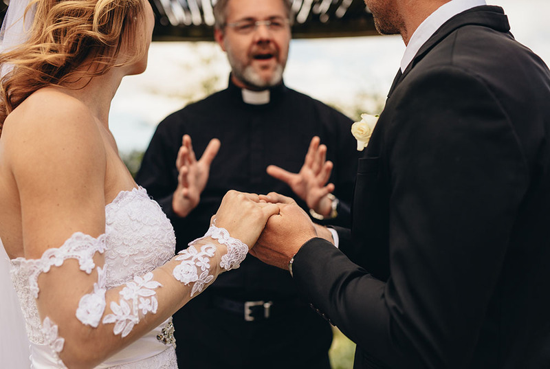 Un ministro oficia una ceremonia de boda para una novia y un novio, la foto muestra a la pareja tomados de la mano y mirando al oficiante mientras extiende las manos y habla durante la ceremonia.