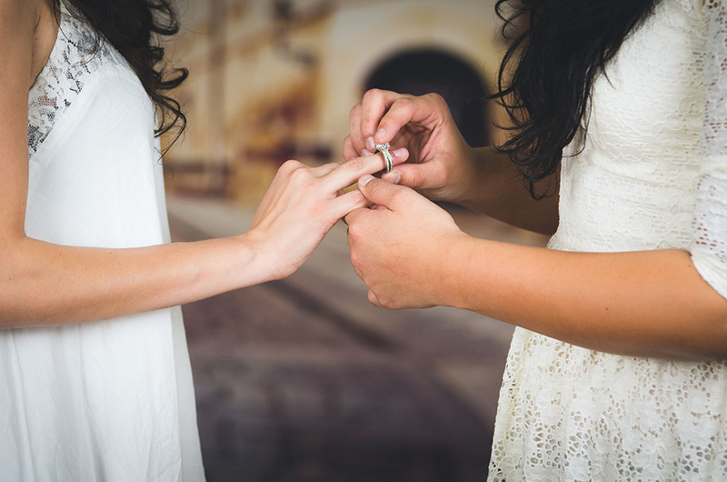 Fotografía de cerca de dos novias con vestidos blancos intercambiando anillos de boda.