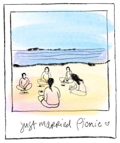 A group of friends and the newlyweds sit in a circle on the beach eating a picnic after a beach wedding