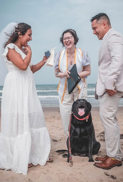 bride and groom stand with officiant on the beach, joined by cute family dog