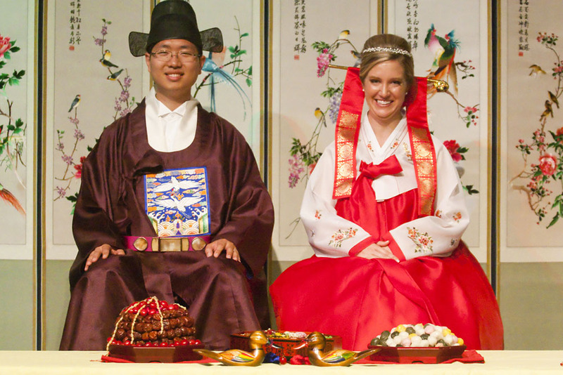 bride and groom pose in traditional korean wedding clothes in front of symbolic food and wooden wedding ducks