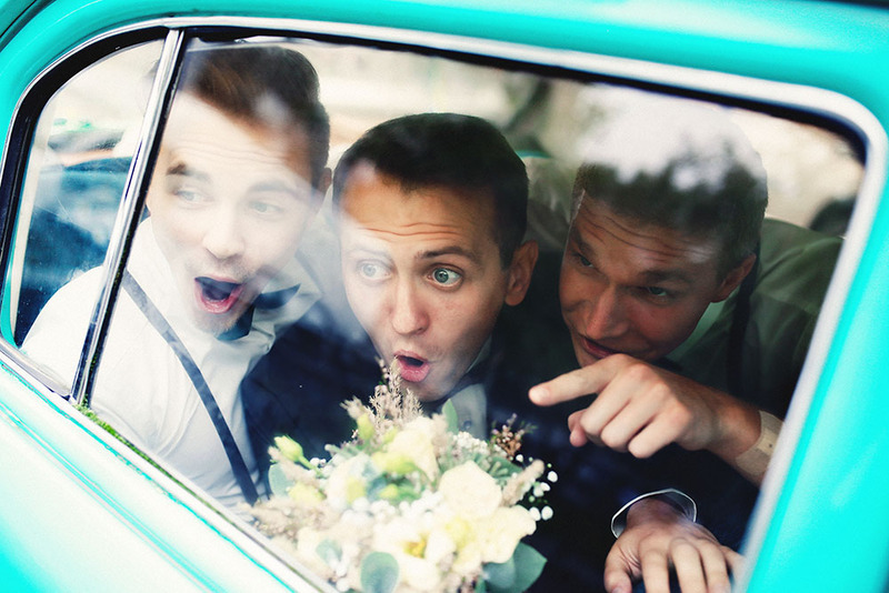 Un novio y un padrino de boda señalan desde la ventanilla de un coche haciendo muecas divertidas el día de la boda.