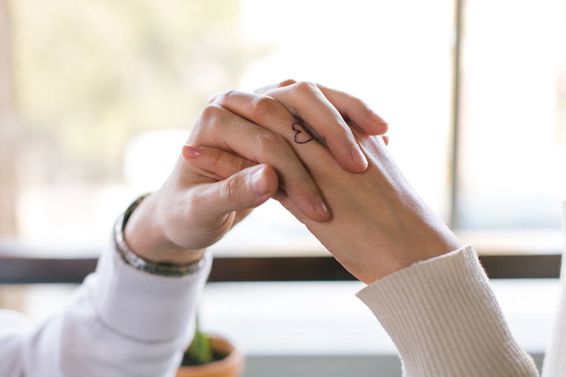 two women hold hands, one with a heart tattoo on ring finger