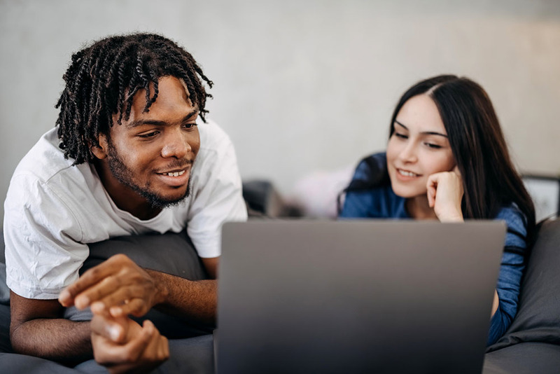 Engaged couple laying on bed, sharing laptop to find wedding vendors