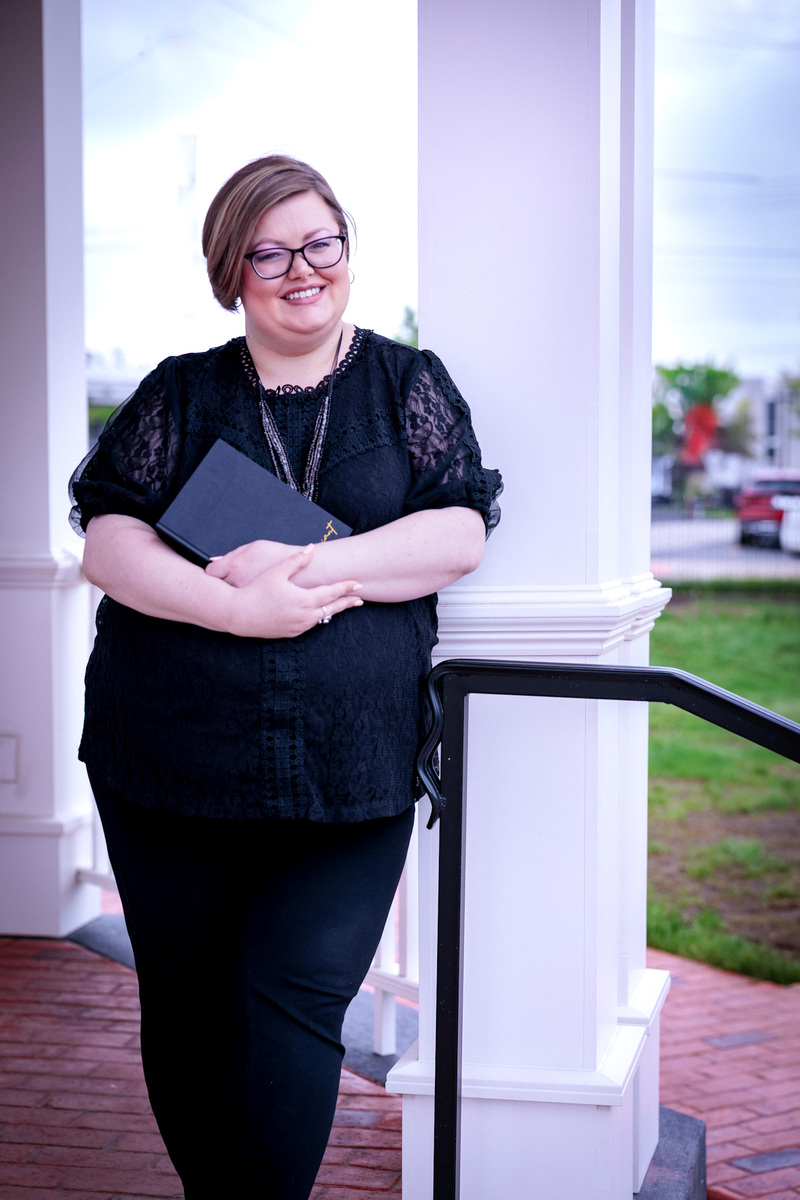 Officiant Brittany Wasson poses with a ceremony binder, smiles at the camera. She is wearing a stylish black outfit and glasses. 