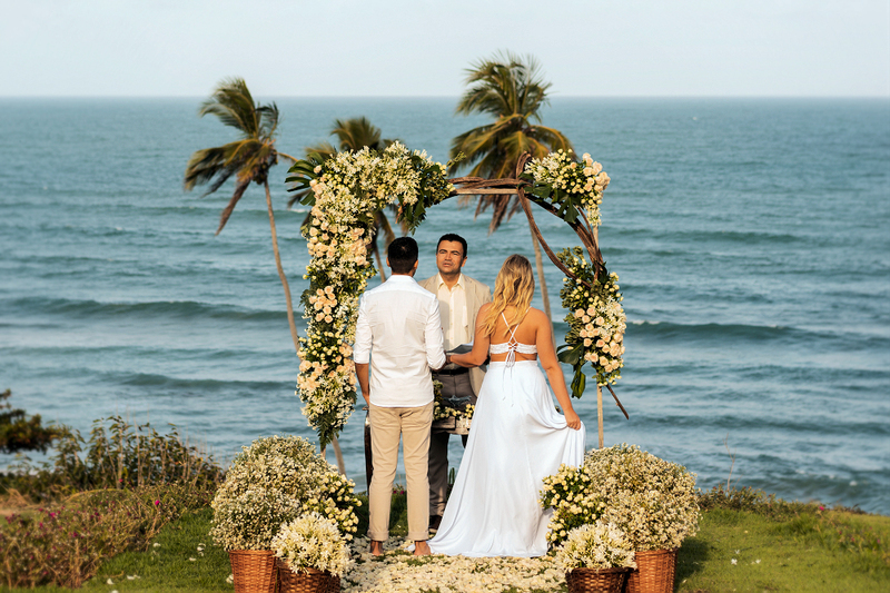 Un oficiante matrimonial realiza una boda frente al océano en la playa con palmeras detrás de él mientras la pareja dice sus votos.
