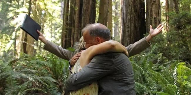 Couple hugs as officiant holds out his arms during wedding in Redwood National Park