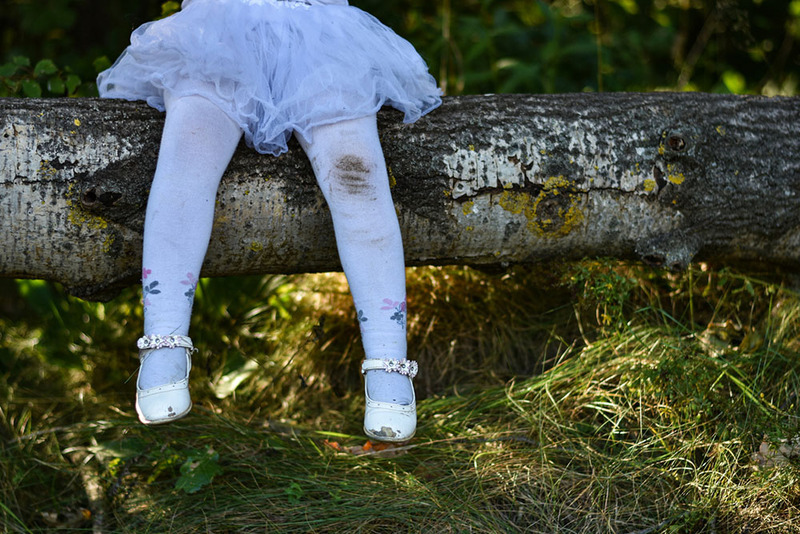 En primer plano se ve a una niña con un vestido blanco y medias sentada en el tronco de un árbol. Tiene una mancha de tierra en la rodilla de las medias.