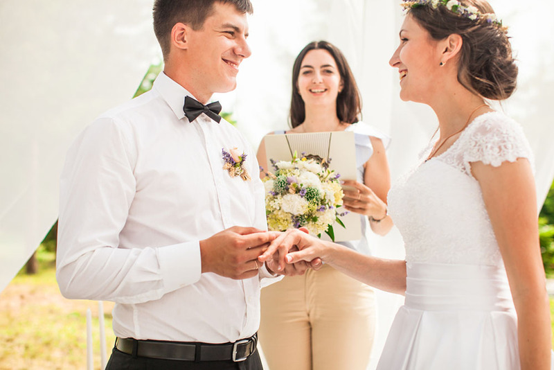 young groom puts ring on bride's finger while wedding officiant smiles.