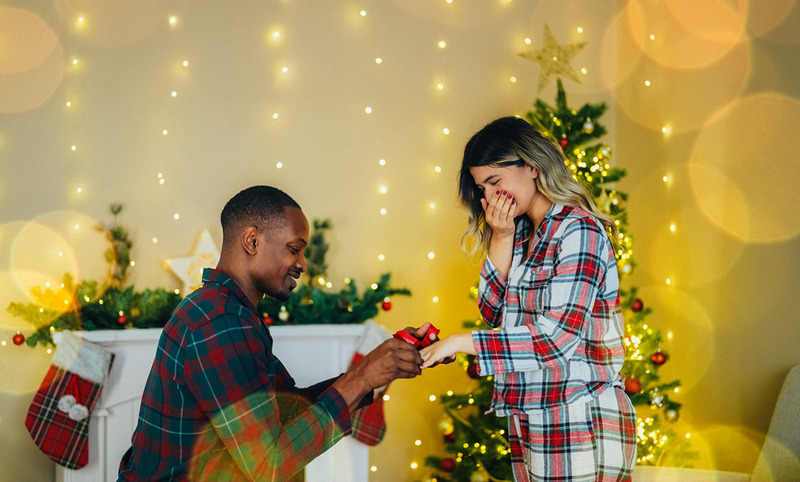 Christmas proposal, man on one knee proposing to girlfriend in front of christmas decorations
