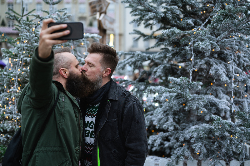 Two men kiss and take a selfie in front of christmas trees