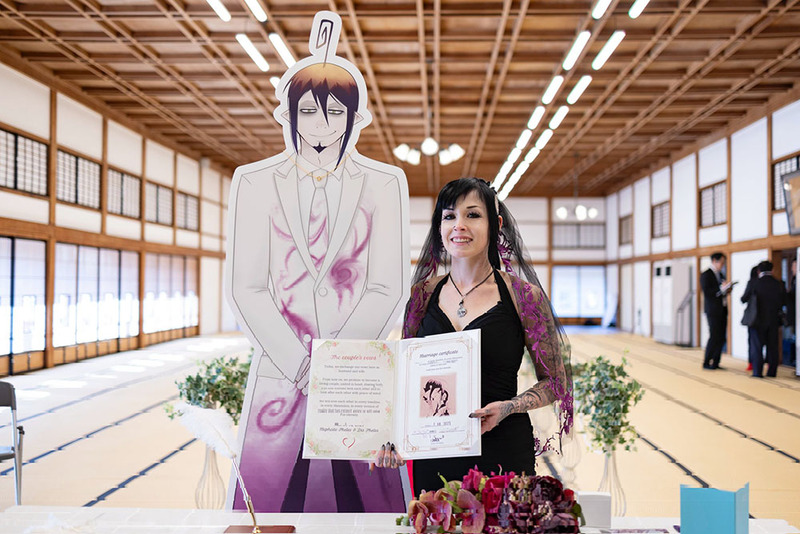 A young bride in a black wedding dress holds a decorative marriage certificate next to a cutout of her anime '2D love'