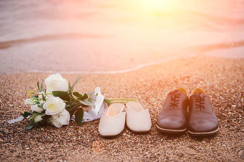 Un ramo de novia y un par de zapatos de boda —para él y para ella— sobre la arena, a orillas del mar al atardecer, durante una ceremonia de boda en la playa.