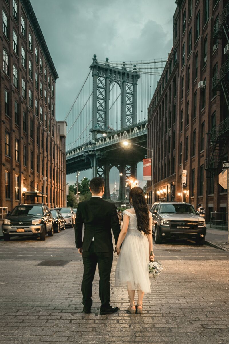couple in wedding clothing stand holding hands, back to camera, facing brooklyn bridge nyc