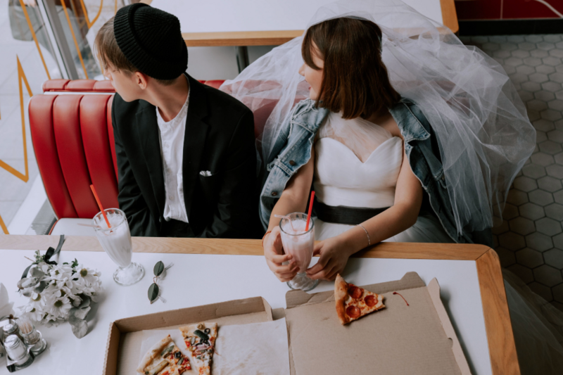 Young couple in modern wedding attire eat pizza in a dinner booth