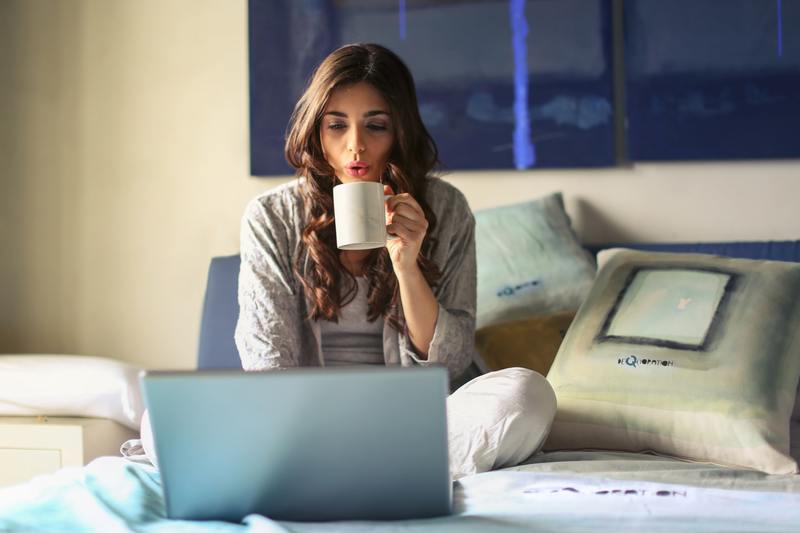 image is a photograph of a woman sitting on a bed, legs crossed and holding a cup of coffee, while she reviews her wedding ceremony script on her laptop before the wedding