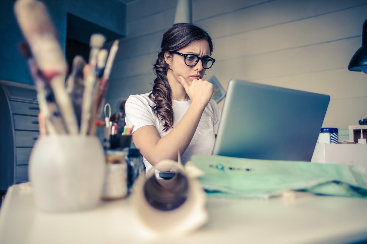 A young woman looks at her laptop computer, resting her chin in her hands and contemplating the best place to get ordained online to officiate a wedding for friends.