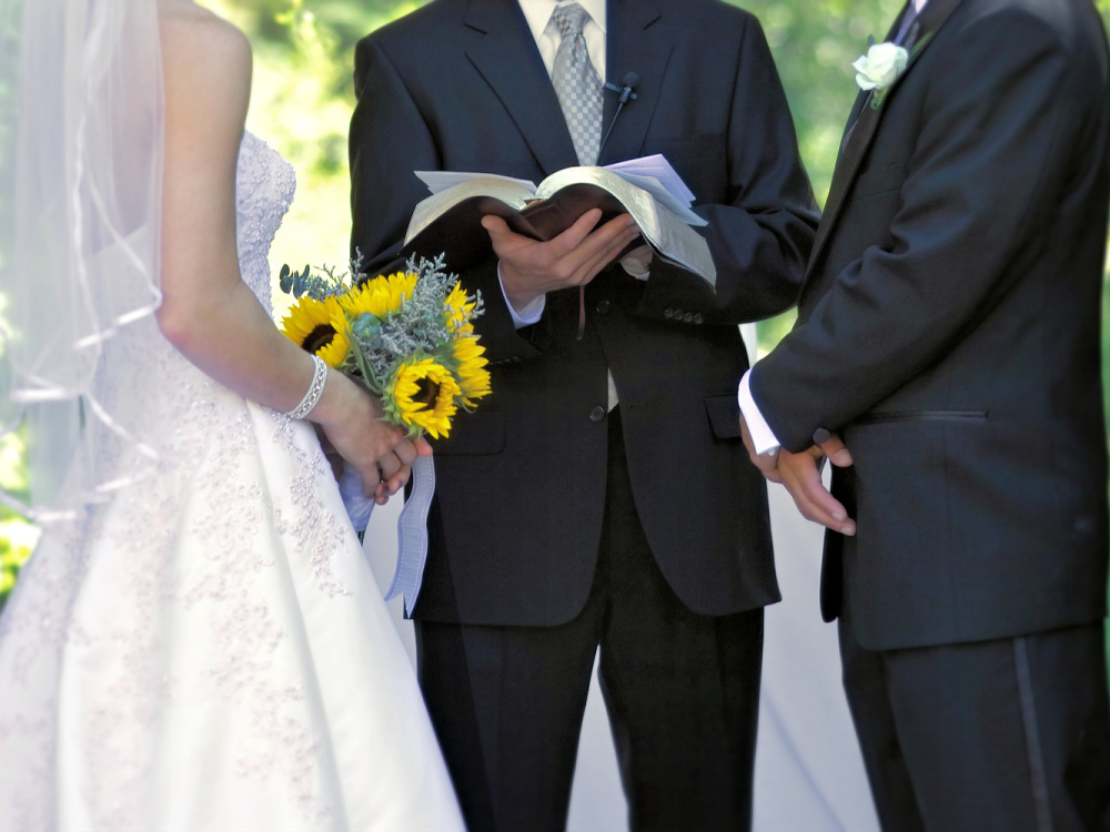 Close up view of bride holding sunflowers and wearing a white dress, next to and officiant reading from a book and the wedding ceremony script, and the groom, wearing a dark suit and a white flower in his lapel. They are outside during the ceremony.