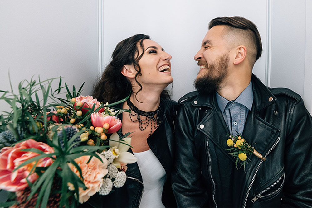 A laughing bride in a white dress and leather jacket poses with the groom, also in a leather jacket and wearing a light blue button up shirt. There is a large wedding bouquet beside them, with pink and orange flowers. They look young and alternative, hip modern couple.