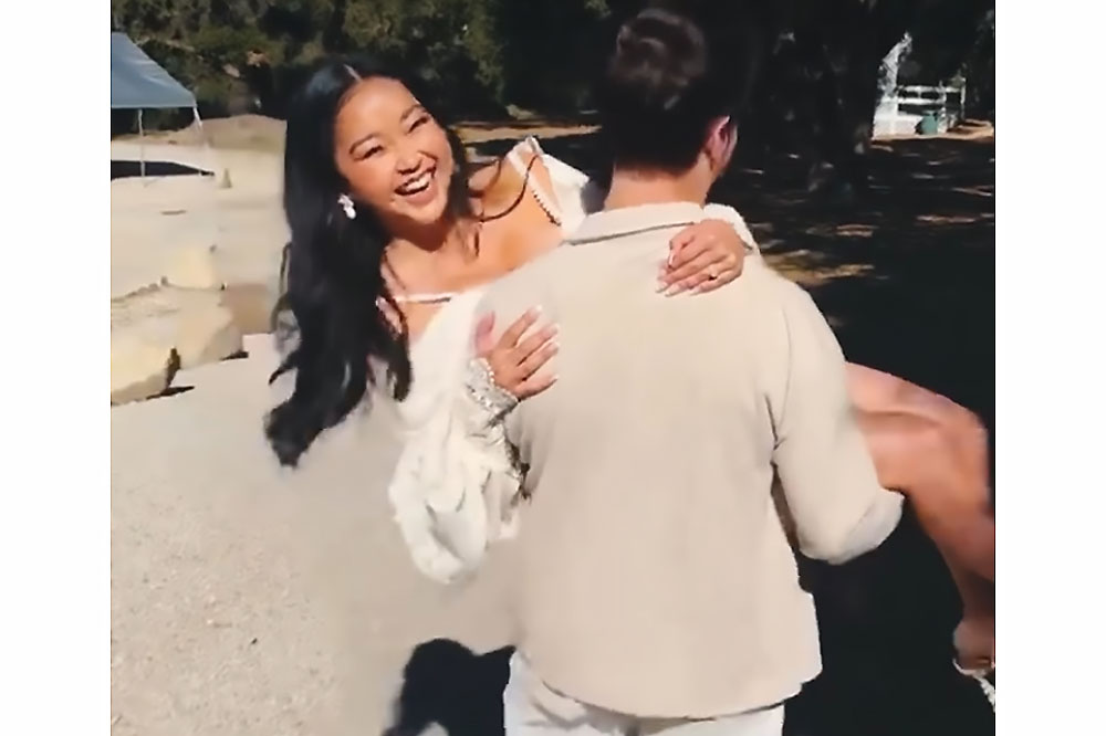 Lana Condor laughs and smiles toward the camera as now-husband Anthony de la Torre carries her in his arms playfully, his back is toward the camera