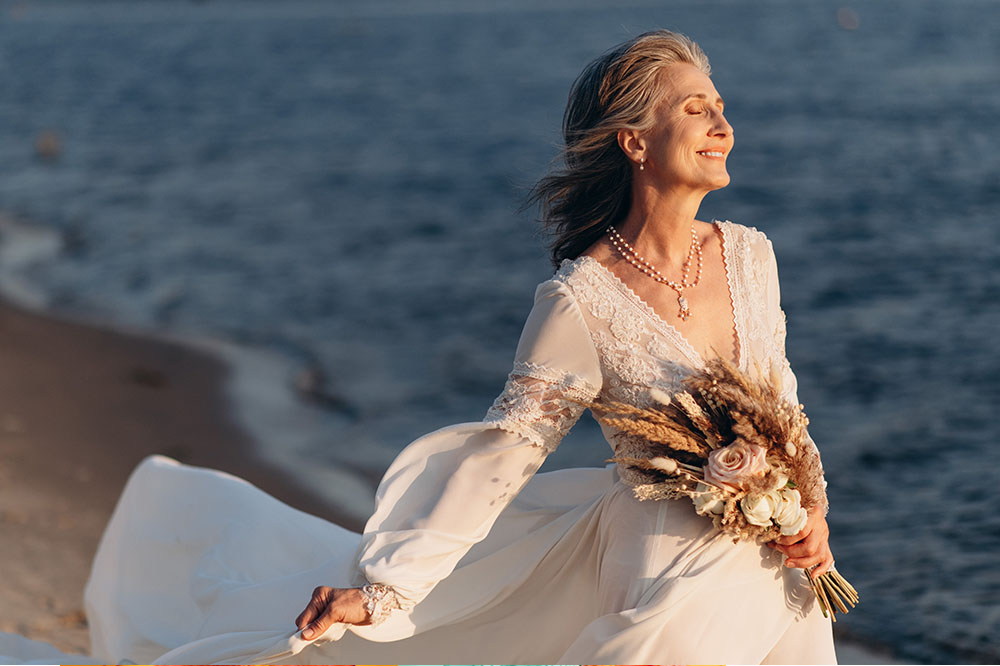 Older bride stands on the beach in a beautiful white wedding dress holding a bouquet of flowers and smiling with her eyes closed