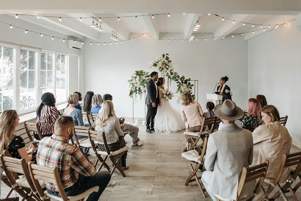 Bride and groom stand with wedding officiant while seated friends and family watch the marriage ceremony