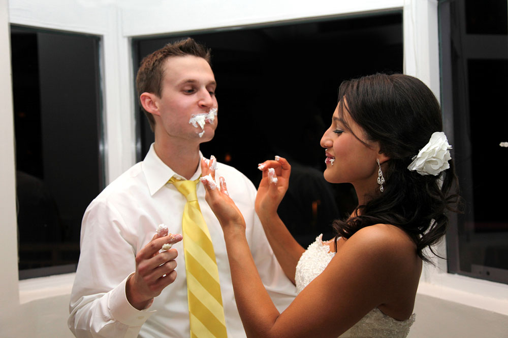 Bride and groom stand together during cake cutting, groom has cake all over his face