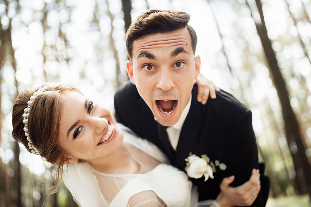 El novio hace una mueca divertida mientras abraza a la novia, posando para fotos de boda al aire libre.
