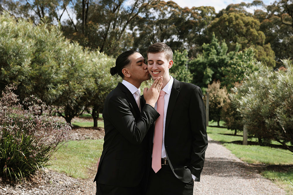 Two young men pose together happily on their wedding day, one kisses the other's cheek