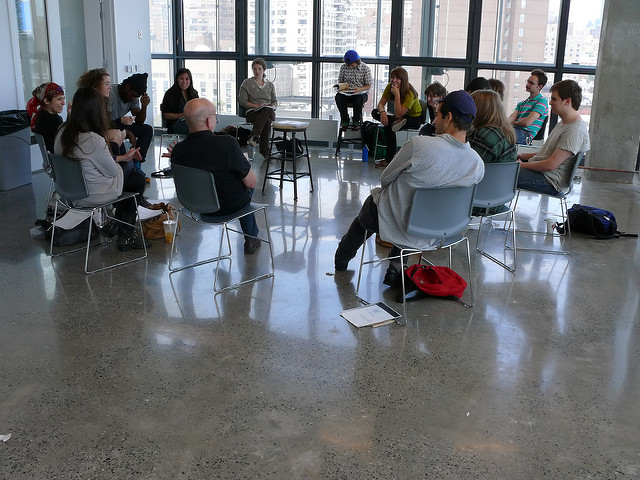 Photo of a group of people sitting in a circle of chairs. Many are leaned back in a casual pose.