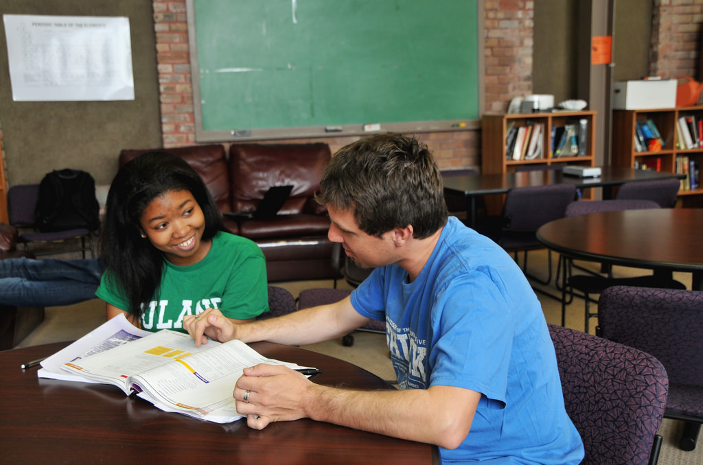 A female student gets help from a male teacher. They're seated at a table in a large classroom, she's smiling, and he's pointing to something in a textbook.