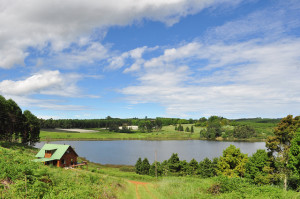 Photo looking down a hill at a lake in the distance, surrounded by one log house with a tin roof and green rolling hills and trees.  More than half the image is blue sky with streaks of white clouds.