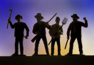 Close-up photo of four plastic figurines, in the shape of men holding axes, pitchforks, and muskets.  Figures are in silhouette against a sunset backdrop