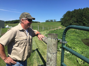 Man in a baseball hat leaning on a fence, looking over green pasture to the right of the camera.