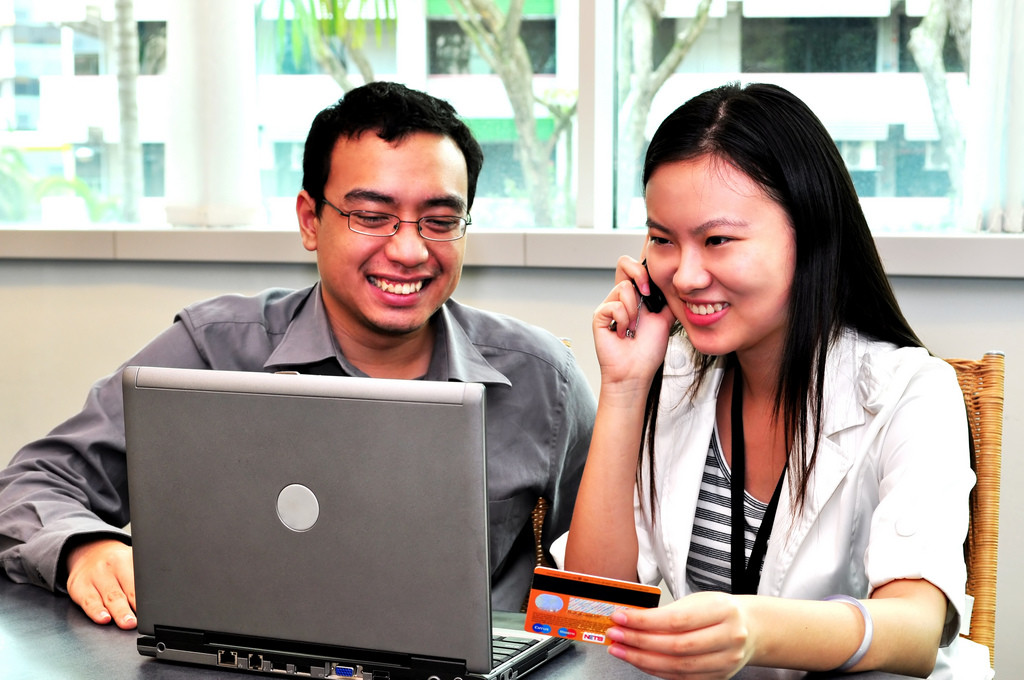 Photo of a man and woman looking at a computer, smiling. The woman is talking on the phone and holding a credit card.