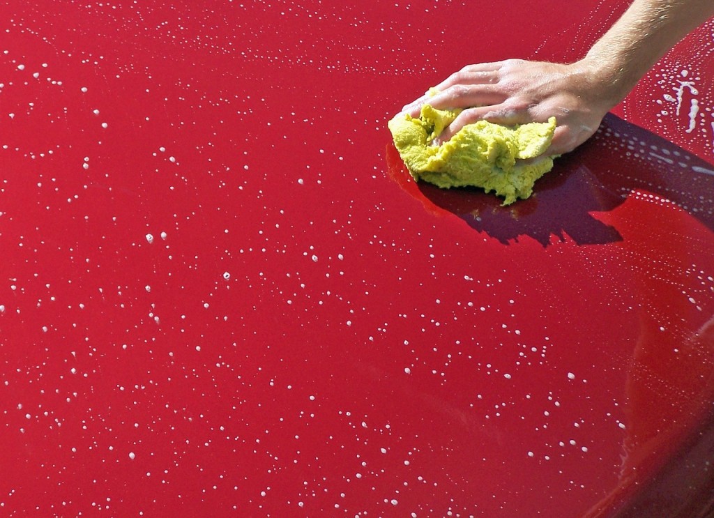 Photo showing a hand holding a yellow sponge, washing the hood of a red car.