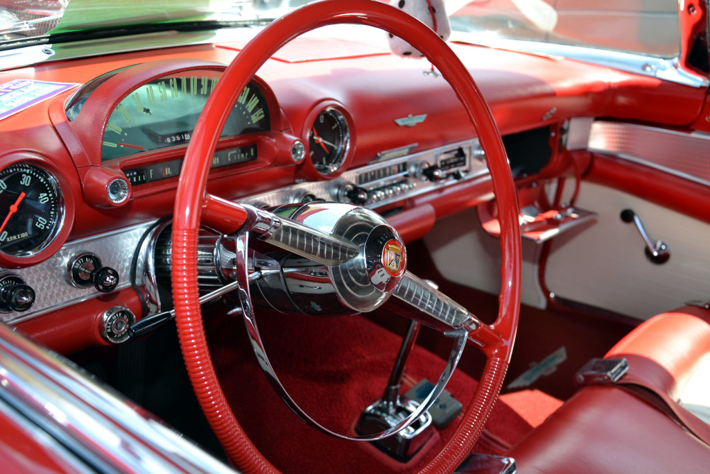 Photo of a cherry red and chrome detailed steering wheel and dashboard of a classic automobile.