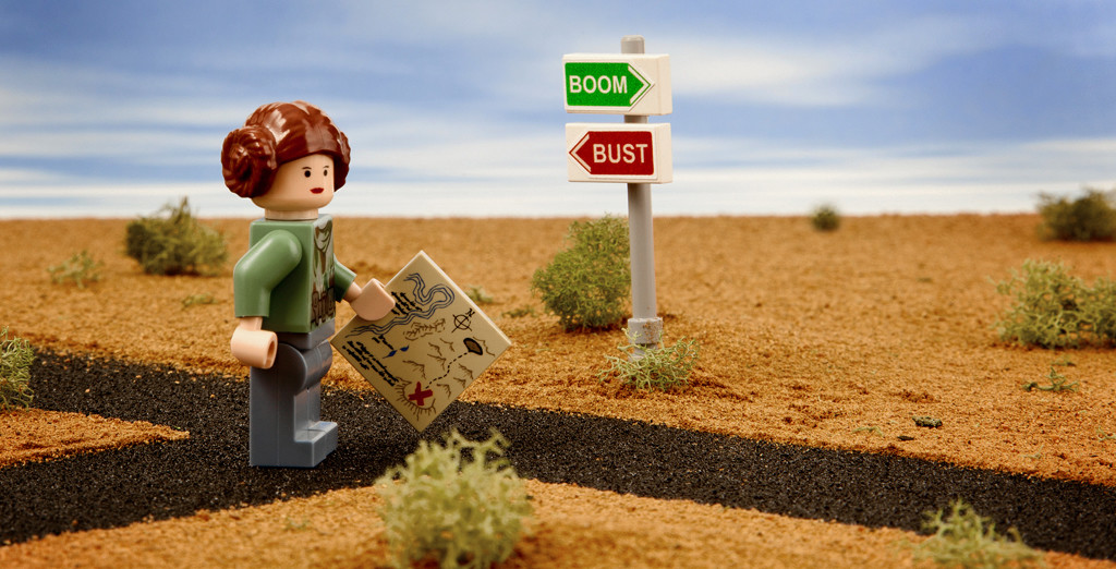 Photo of a Lego woman holding a map, standing at a crossroads labeled "Boom" and "Bust."