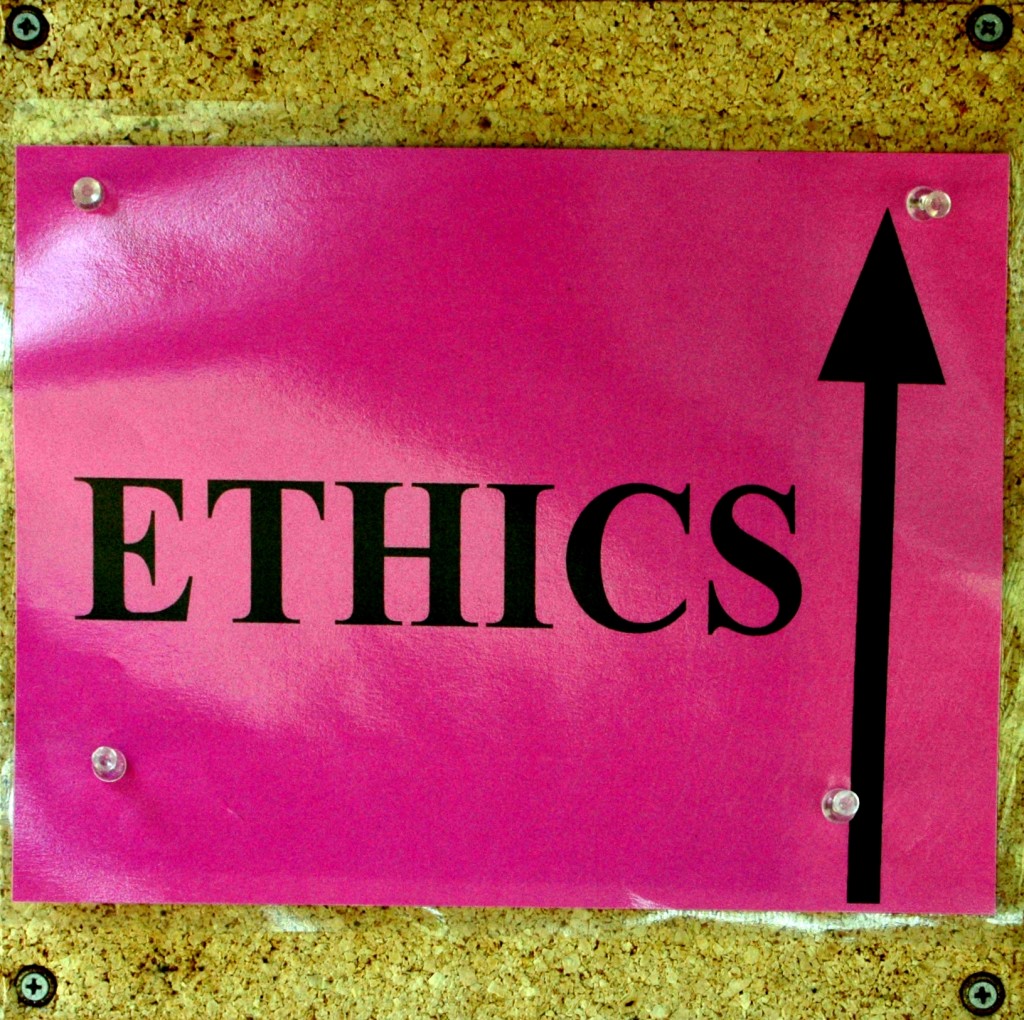 Bright pink laminated paper thumbtacked to a cork board. The pink sign contains the word ETHICS in black; to the right of the word is a black arrow pointing up. 