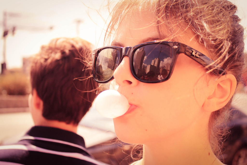 Teenage girl in foreground wearing sunglasses, blowing a gum bubble. Boy in background facing the opposite direction.