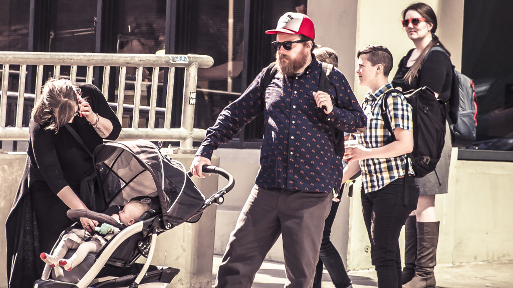 Un homme barbu portant un chapeau rouge pousse une poussette pendant qu'une femme surveille l'enfant et parle sur son téléphone portable. Deux jeunes gens en arrière-plan. Des hipsters de Seattle.